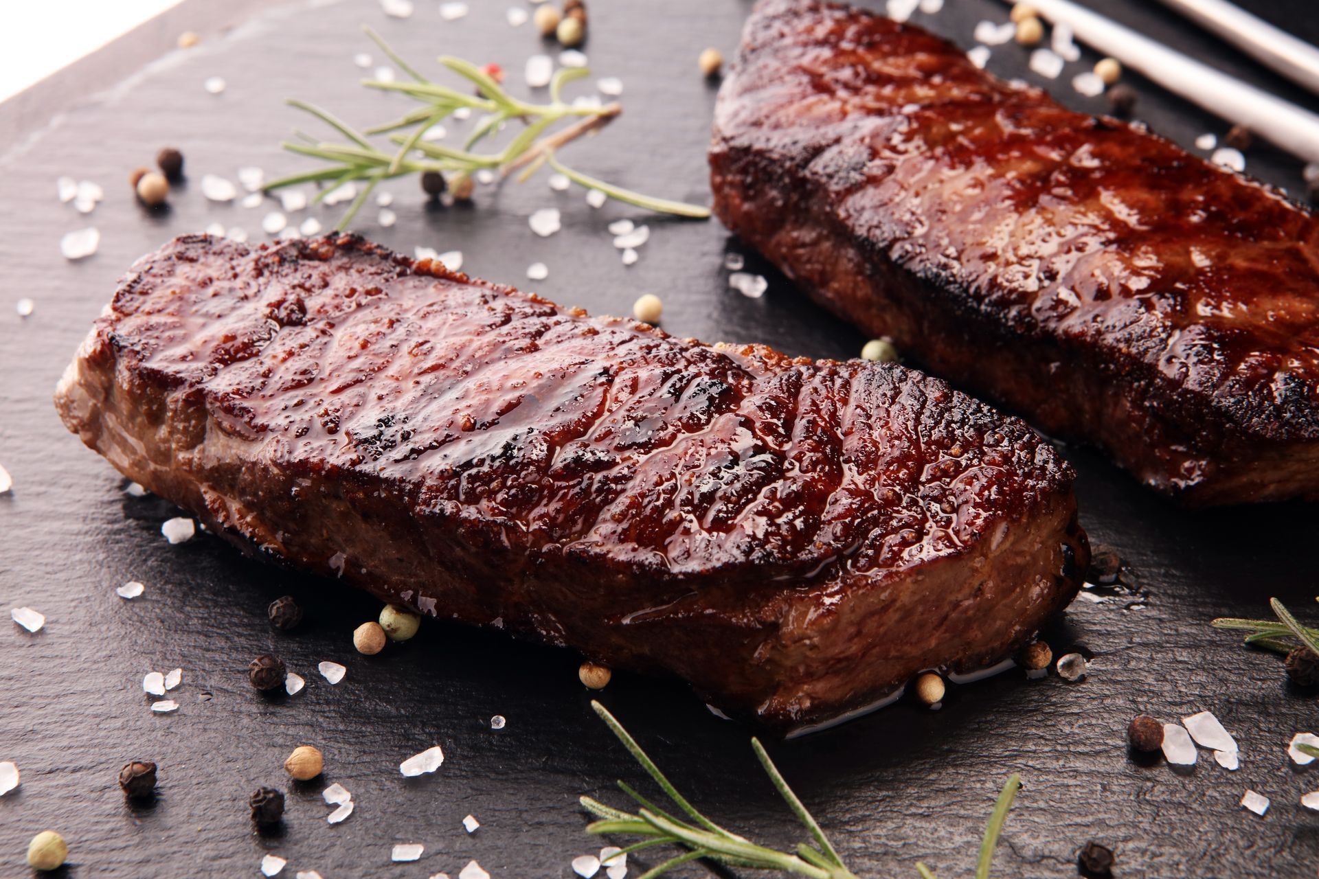 Two cooked steaks on a black surface, sprinkled with salt, pepper, and rosemary.