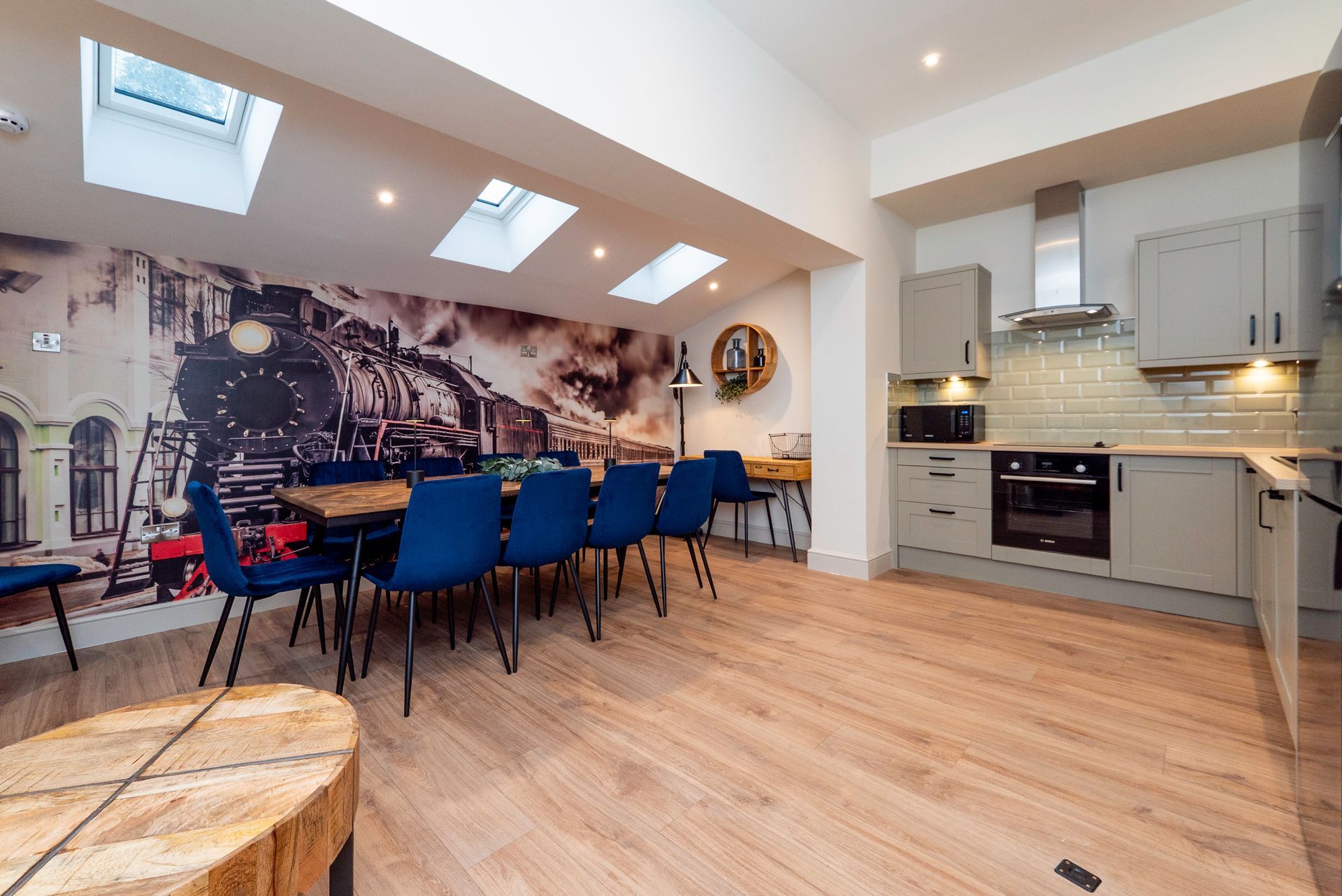 Open-plan kitchen and dining area with a train mural and a long table with blue chairs.