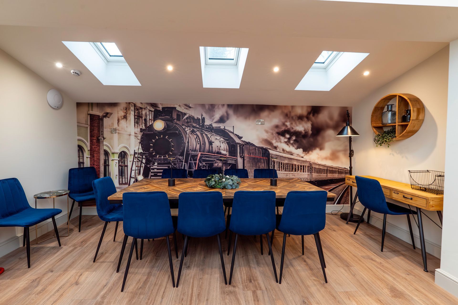 Dining room with a long table, blue chairs, a train mural, and skylights.