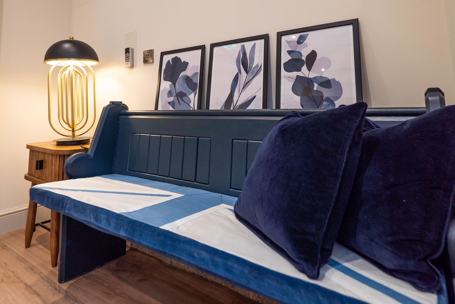 Blue bench with velvet cushions, artwork, and lamp on a wood floor.