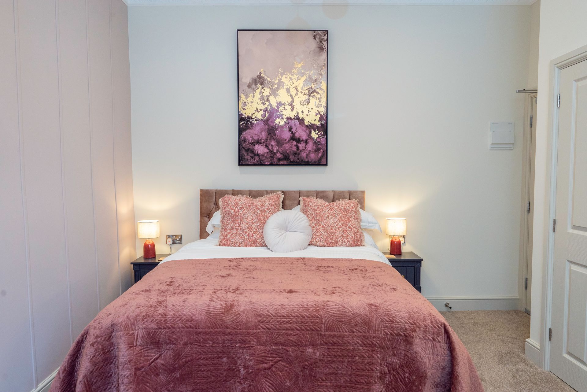 Bedroom with a bed, nightstands, artwork, and a closed door. Walls are white and pink, bedspread is pink.