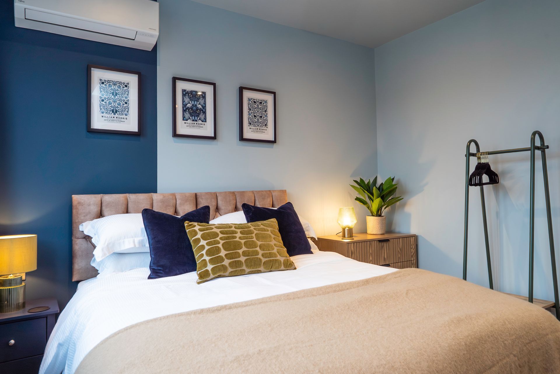 Bedroom with blue and tan walls, a bed with pillows, and artwork. A standing rack is on the right.