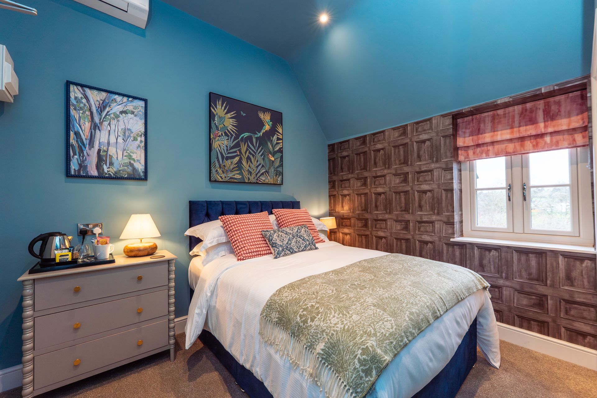 Bedroom with blue walls, double bed, wooden dresser, art, window with red shade.
