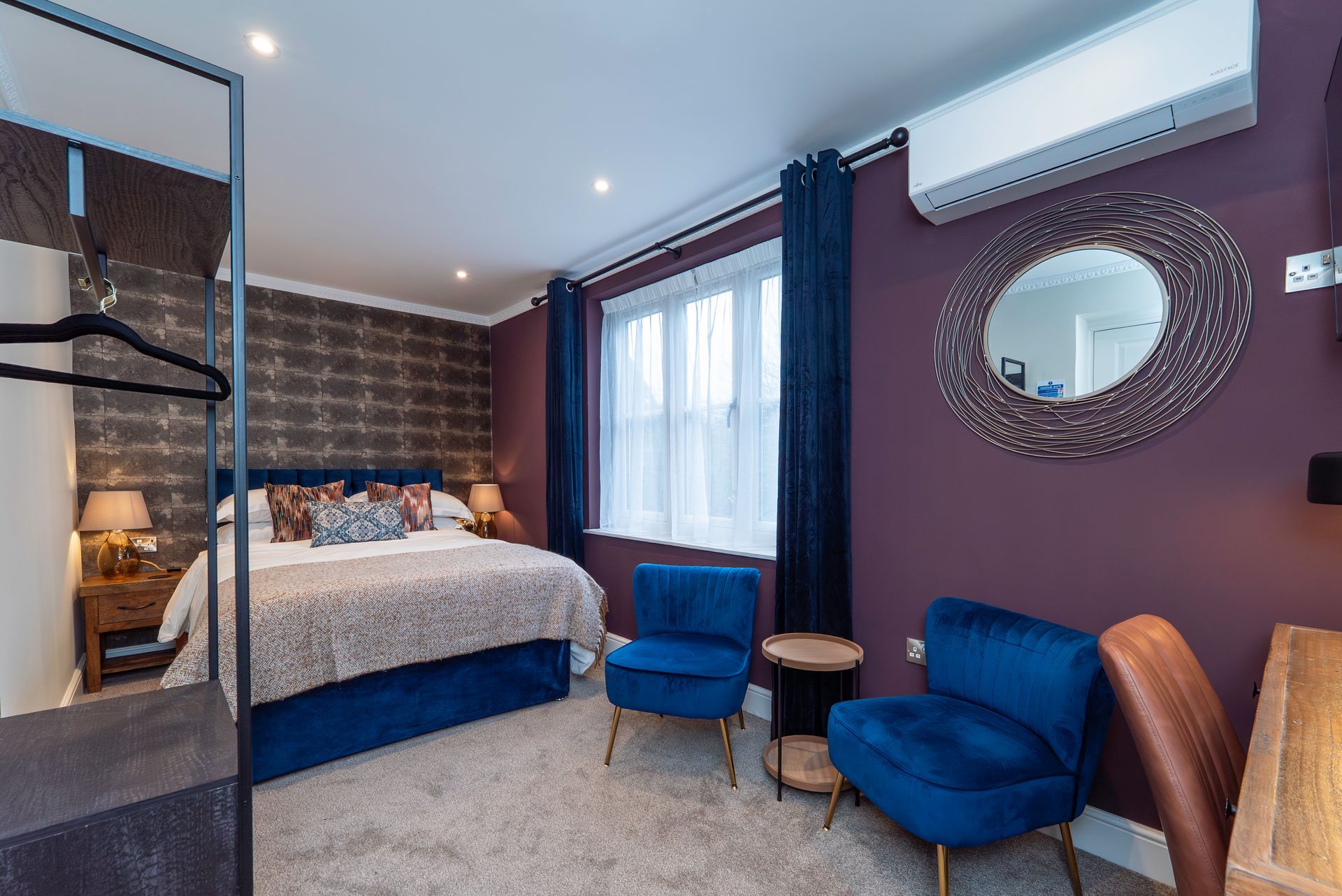 Bedroom with a bed, two blue chairs, and a round mirror on a purple wall.
