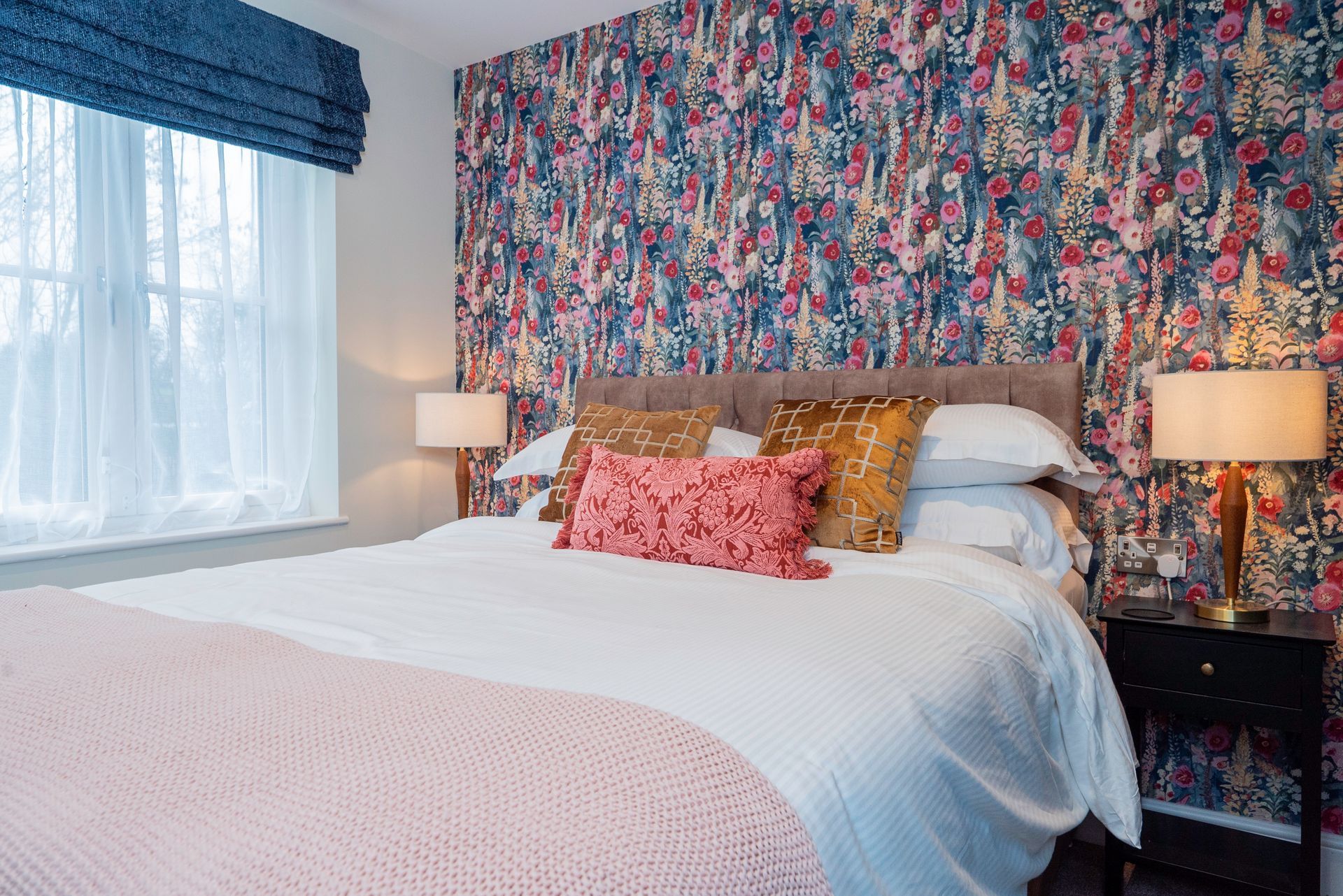 Bedroom with floral wallpaper, bed, pillows, lamps, and a window with a blue shade.