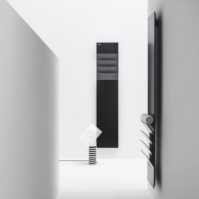 Due radiatori verticali minimalisti, di colore grigio scuro, montati a parete in una stanza bianca e luminosa, uno dei quali dotato di portasciugamani integrato.