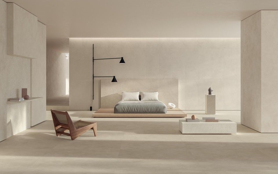 Una camera da letto minimalista con pareti color crema, un letto a piattaforma, un tavolino basso, una poltrona in legno e una lampada da parete doppia.
