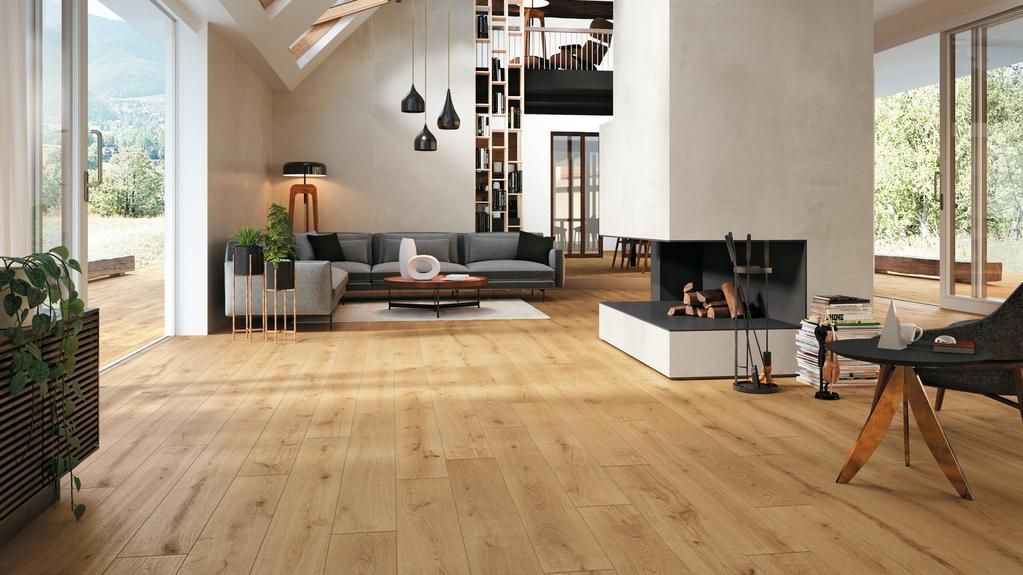 Un ampio e moderno soggiorno con pavimento in legno chiaro, divano angolare grigio, camino e grandi finestre.