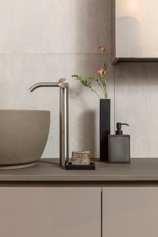Un moderno mobile da bagno con lavabo, rubinetto alto in metallo, saponetta su un vassoio, un vaso sottile con fiori e un dispenser di sapone.