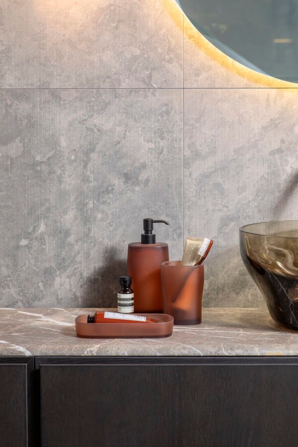 Accessori da bagno color ambra e un set di spazzolini da denti su un piano in pietra grigia, contro una parete rivestita di piastrelle effetto marmo.