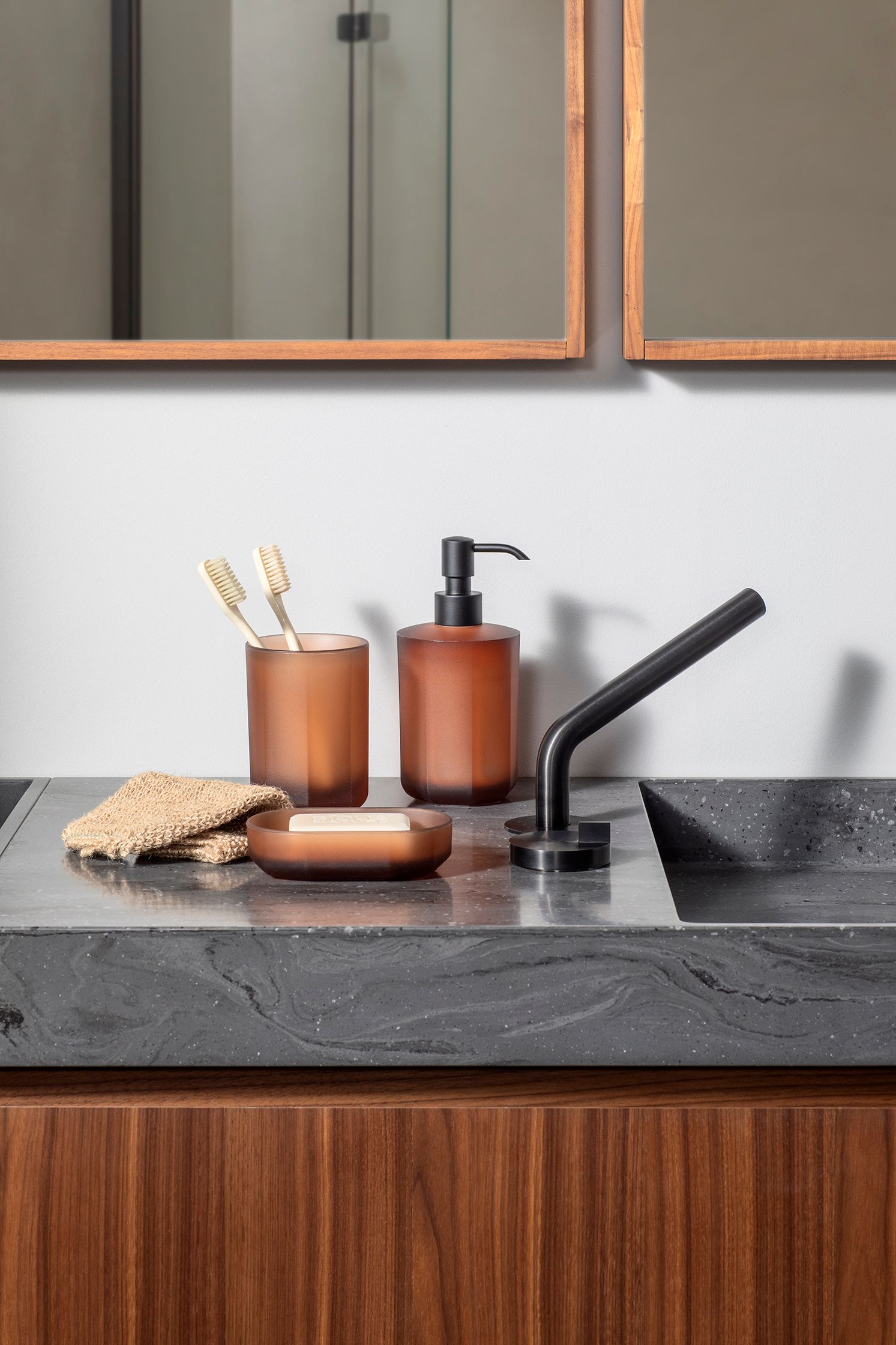 Portaspazzolino, dispenser di sapone e piattino color ambra su un mobile da bagno in cemento grigio scuro con rubinetto nero.