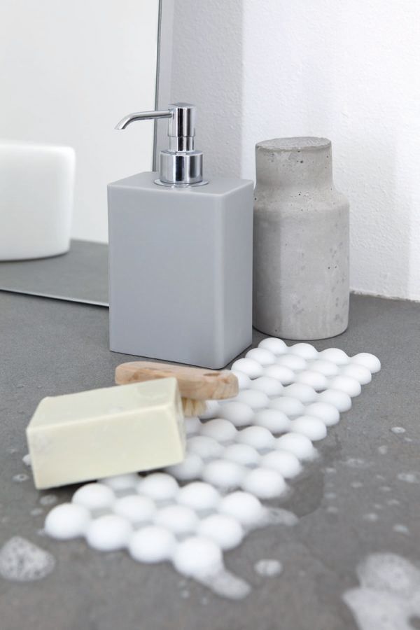 Un dispenser di sapone grigio, un contenitore in cemento e una saponetta appoggiati su un tappetino da bagno bianco, ruvido e dalla superficie irregolare, su un ripiano.