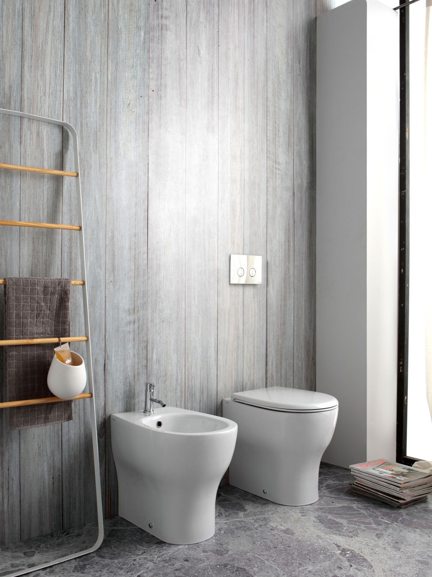 Un bagno moderno con bidet e WC bianchi su una parete rivestita in legno grigio, con una scaletta portasciugamani nelle vicinanze.