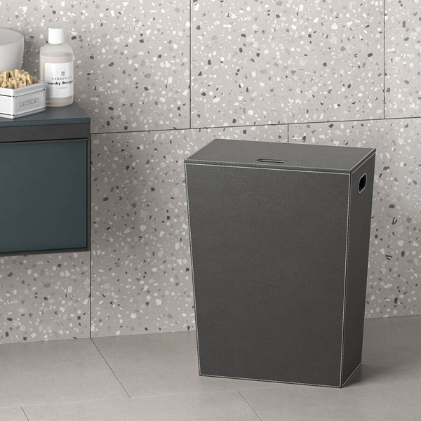 Un alto cesto portabiancheria con coperchio, di colore grigio scuro, è appoggiato su un pavimento chiaro di fronte a una parete rivestita di piastrelle grigie effetto terrazzo.