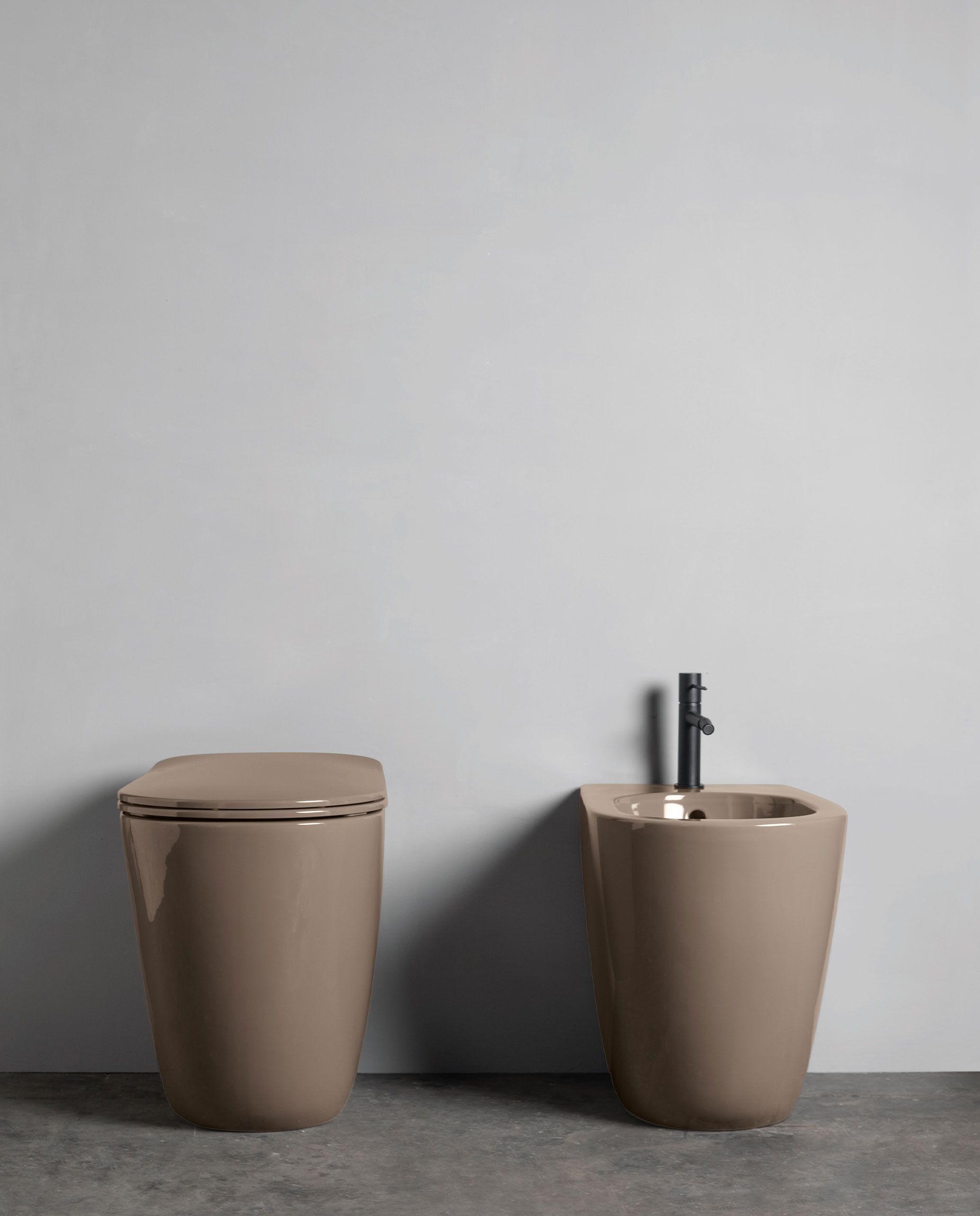 Un WC e un bidet beige a filo muro con rubinetto nero su una semplice parete grigia.