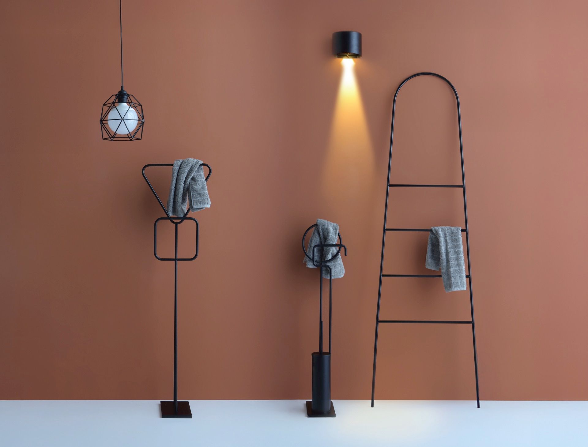 Accessori da bagno moderni in metallo nero, tra cui due portasciugamani, una scaletta e una lampada a sospensione, su una parete marrone.
