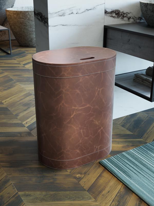 Un cesto portabiancheria alto e ovale in pelle marrone con coperchio, posizionato su un pavimento in legno a spina di pesce in un bagno moderno.