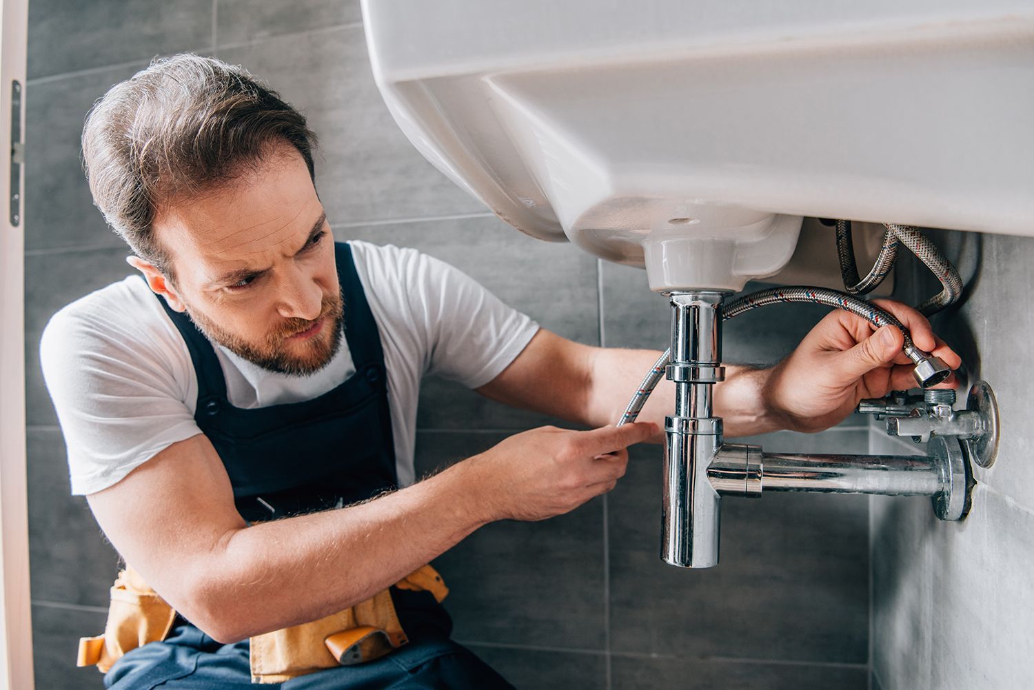 A plumber in working overalls is fixing a sink in the bathroom. A plumber in working overalls is fixing a sink in the bathroom.