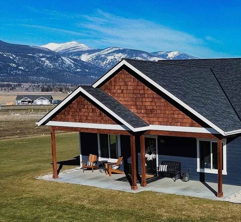 A modern home with dark blue siding, brown shingle accents, and a front porch, set against snow-capped mountains.