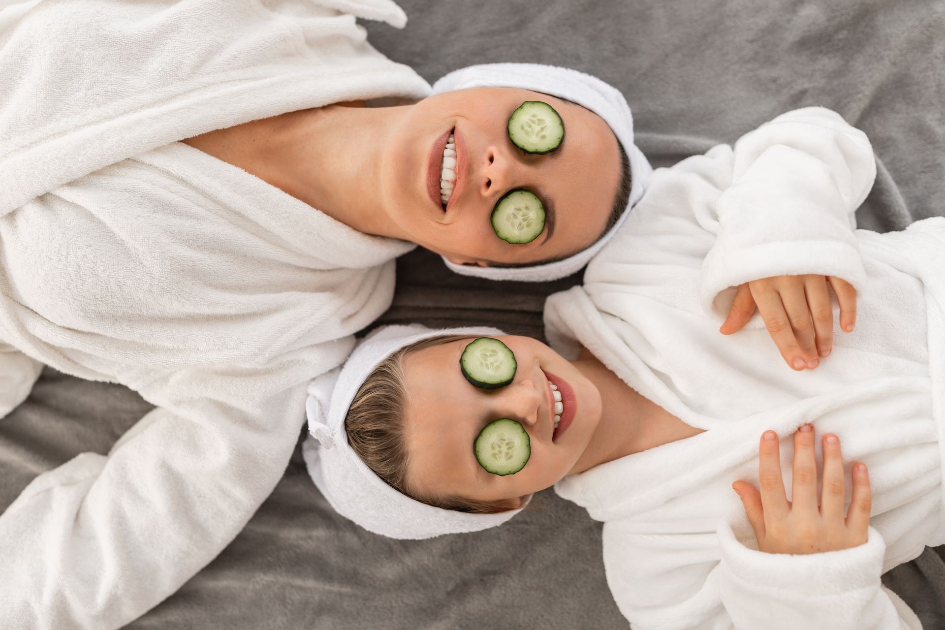 Two people wearing white robes and headbands lie on a gray surface with cucumber slices over their eyes.