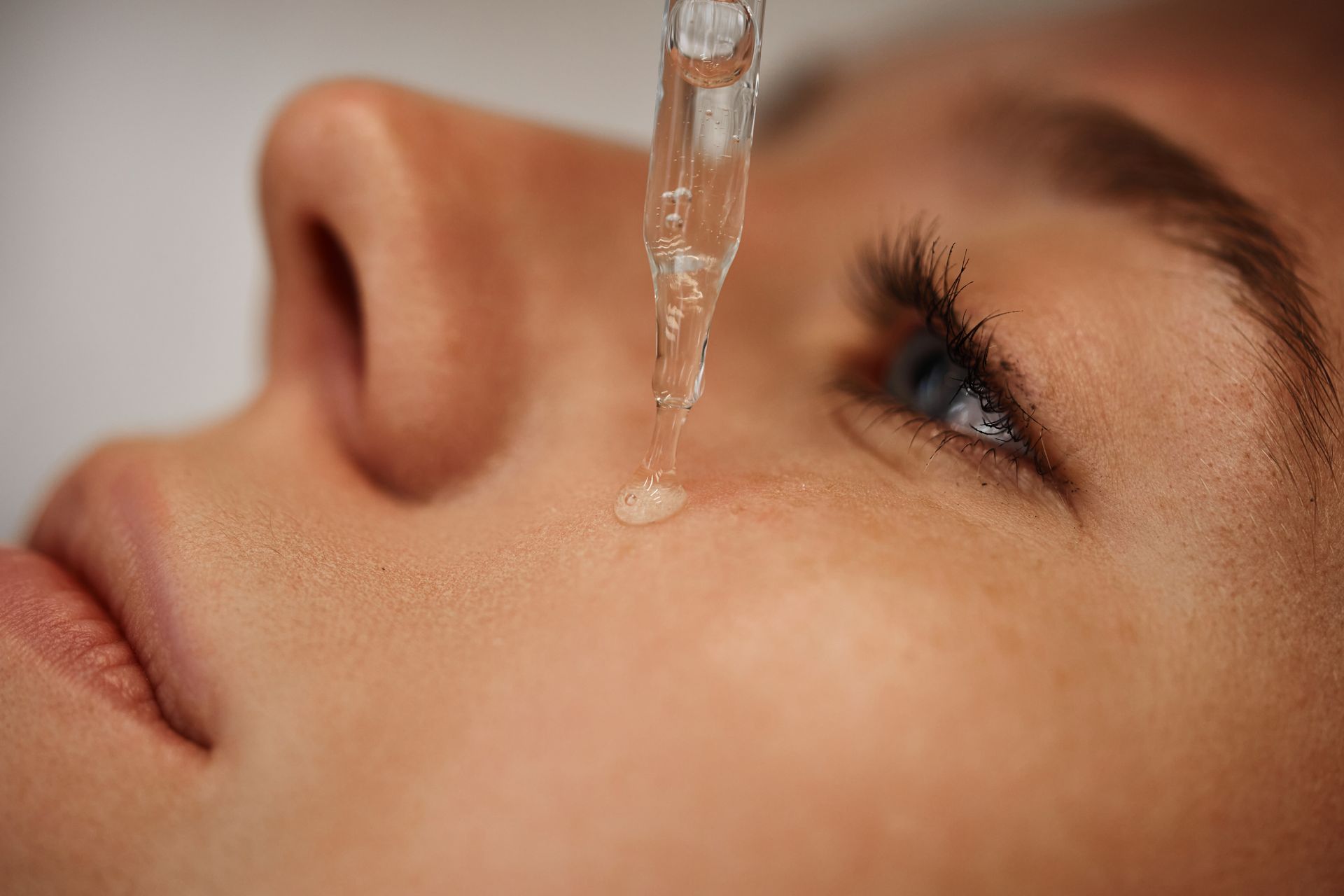 A glass dropper applies a clear liquid serum onto a person's cheek near their eye.
