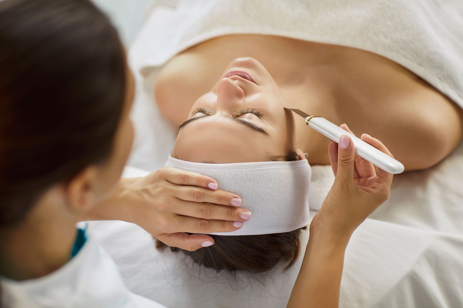 A practitioner uses an ultrasonic skin spatula device on a client's forehead during a facial treatment.