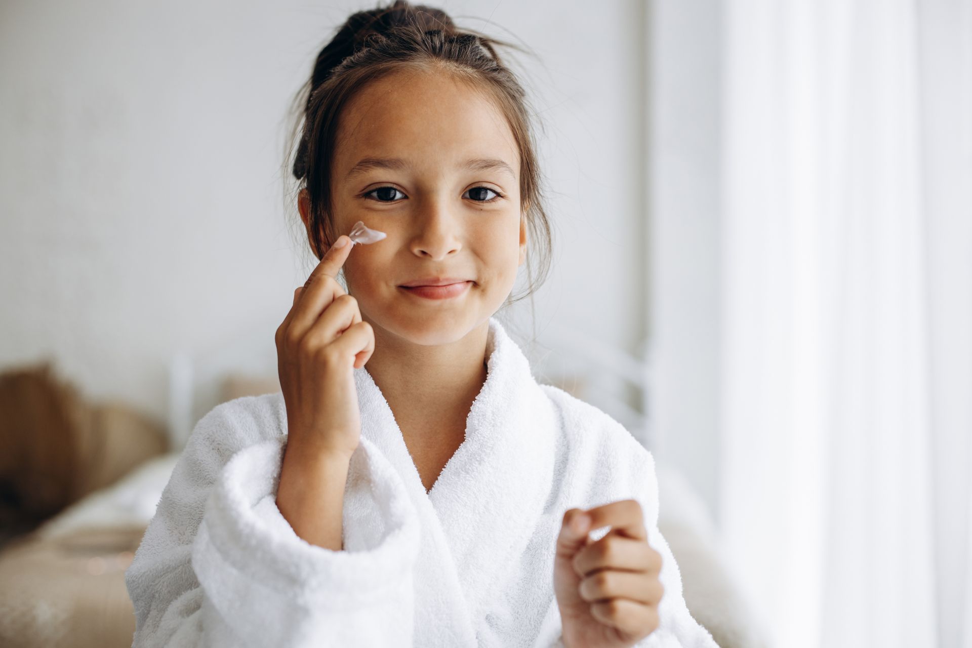 A person in a white bathrobe applies a small piece of clear facial tape to their cheek while smiling.