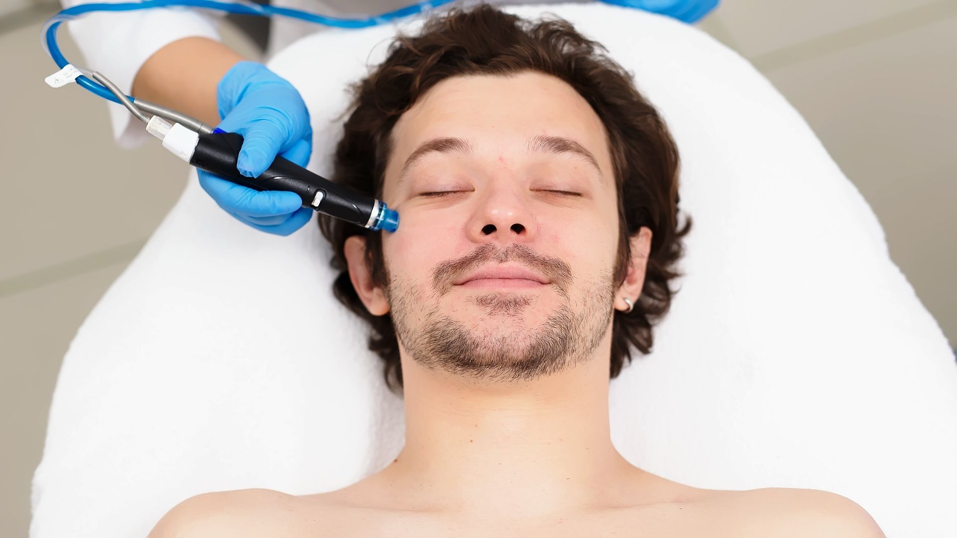 Man receiving facial treatment, blue-gloved hand holding device to his face; white background.