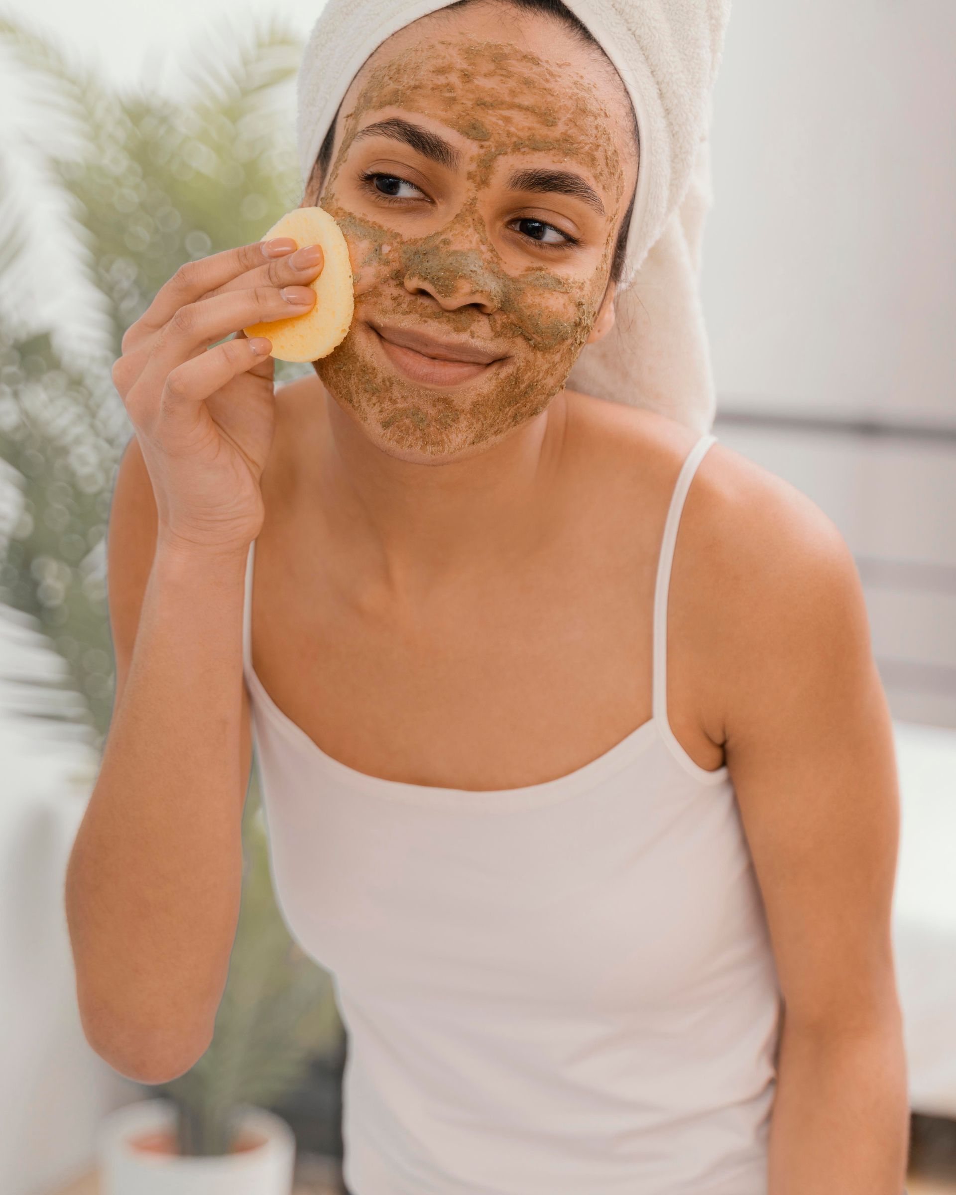 A woman with a towel wrapped around her head is applying a mask to her face.