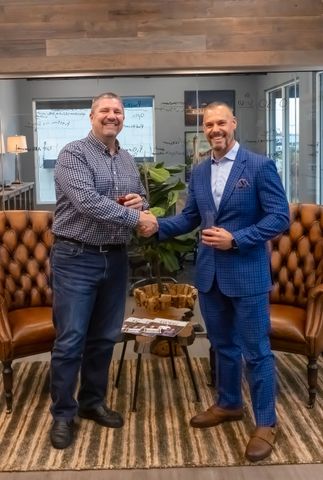 Two men are shaking hands in a living room while holding wine glasses.
