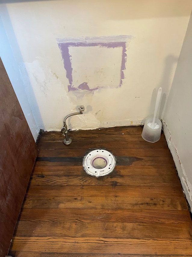 A wooden floor in a bathroom with a toilet flange and a toilet brush.