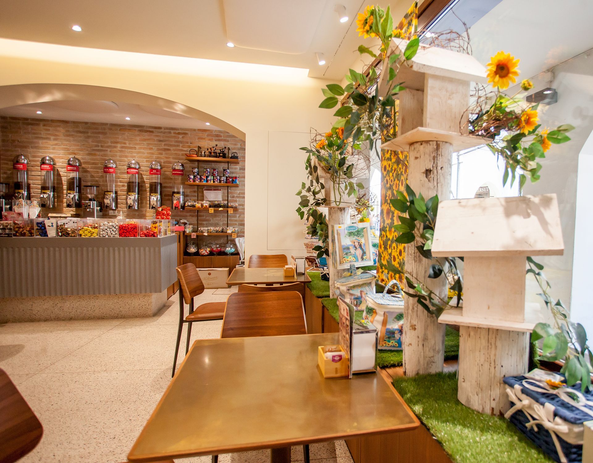Accogliente interno di una caffetteria con tavoli in legno, bancone e decorazioni floreali vicino a un'esposizione ricoperta di piante.