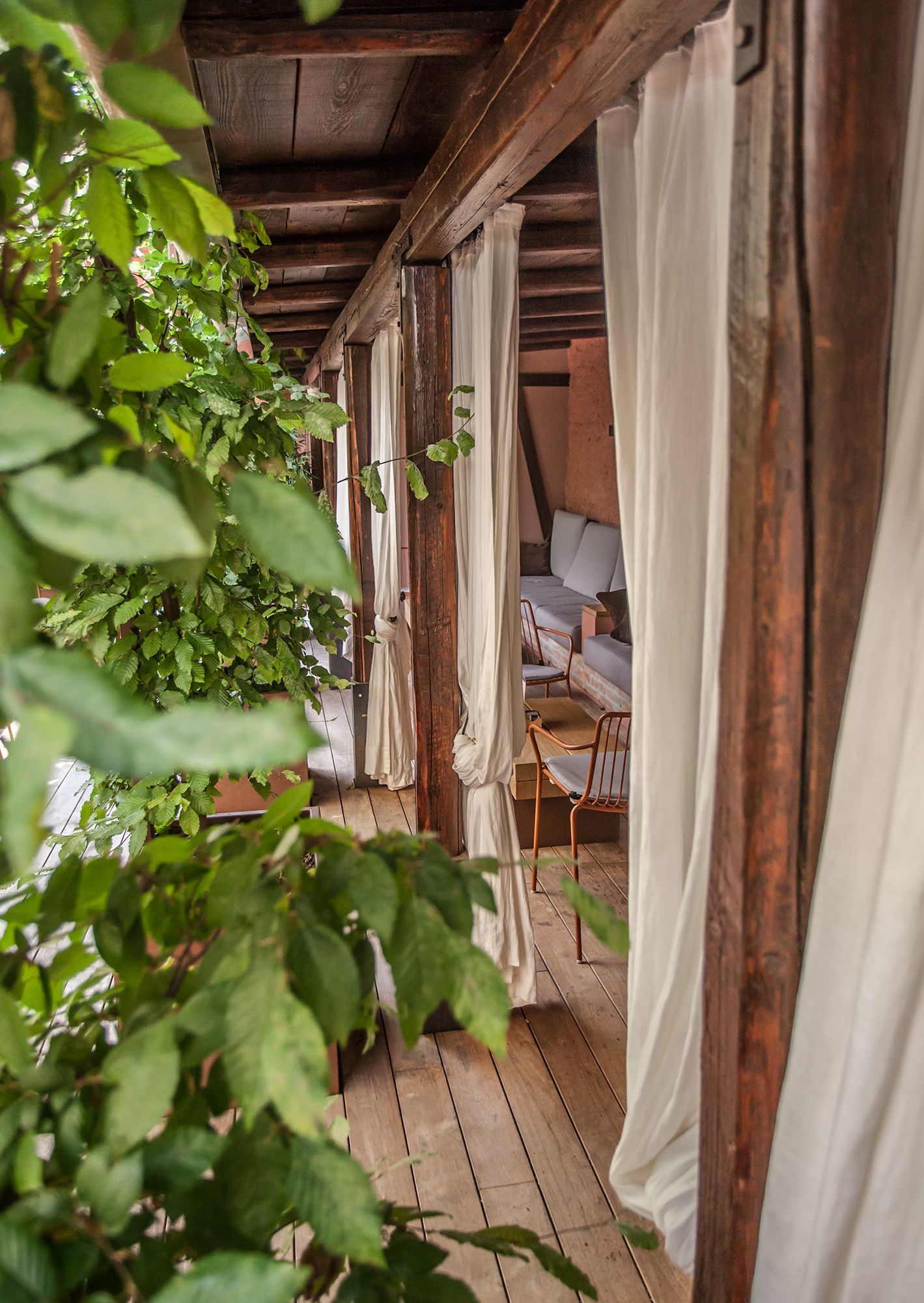 Veranda in legno con piante rampicanti, tende bianche e vista su un interno rustico