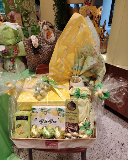 Cesto regalo con carta da regalo gialla, nastro verde e assortimento di dolcetti confezionati, esposto in un negozio.