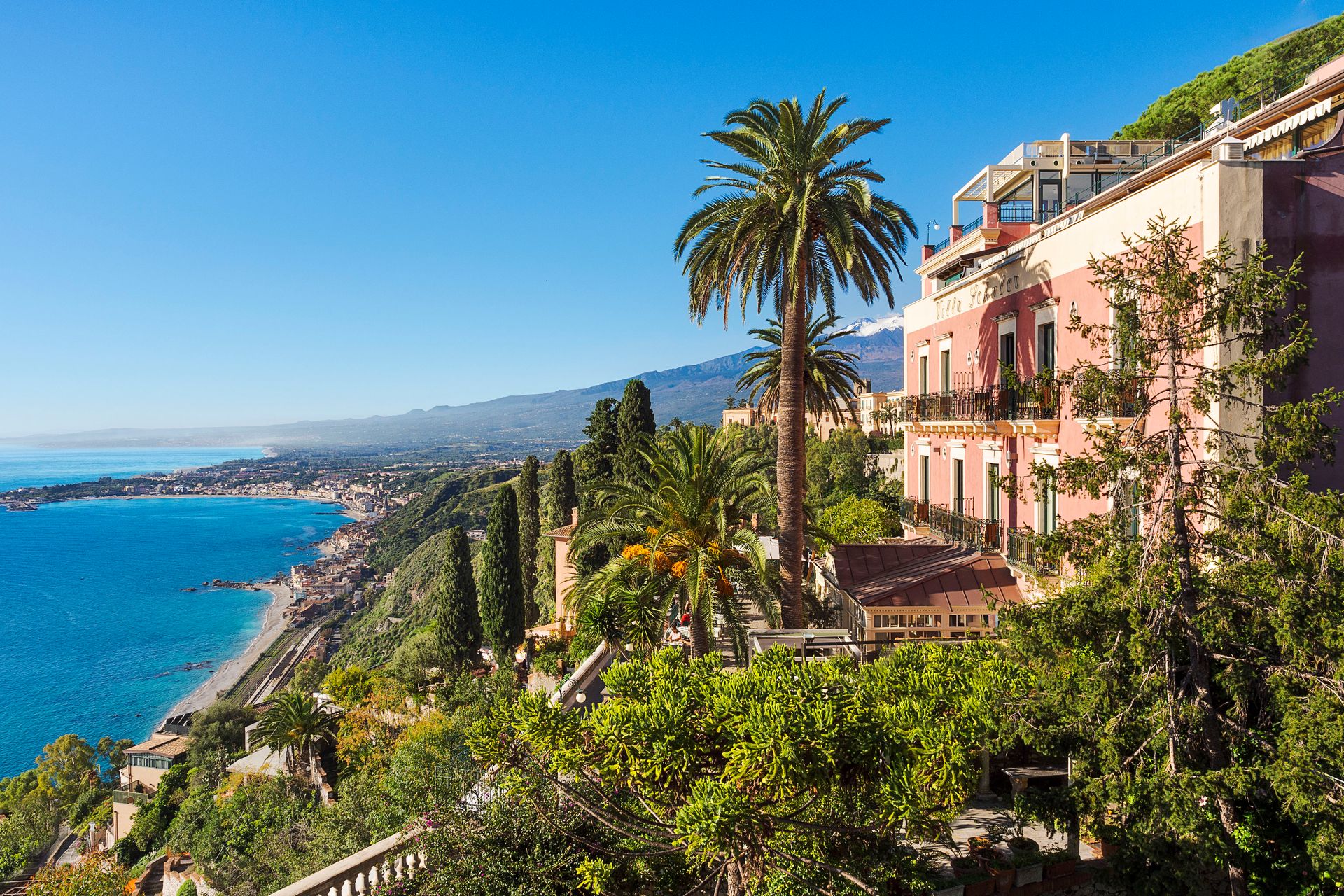 Coastal hillside with pink buildings, palm trees, and blue sea under a clear sky