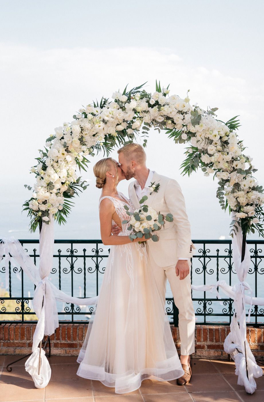 Bride and groom kissing under a white floral wedding arch outdoors