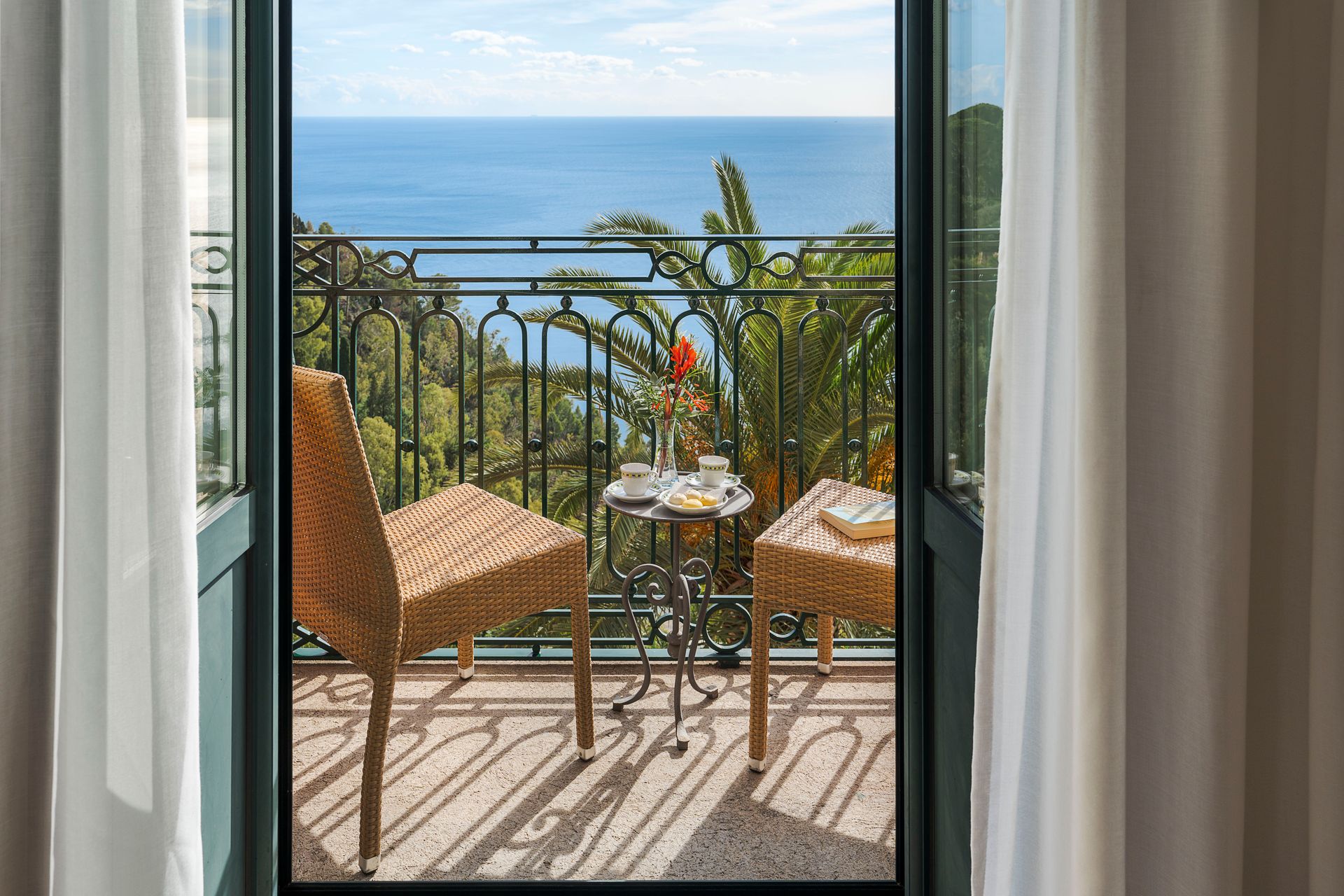 Balcony with wicker chairs and small table overlooking the sea, framed by open curtains