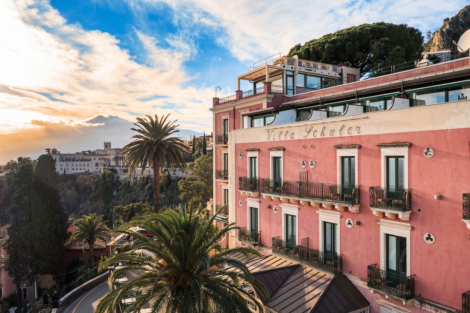 Edificio rosa en la ladera con balcones y palmeras al atardecer con vistas a la costa.
