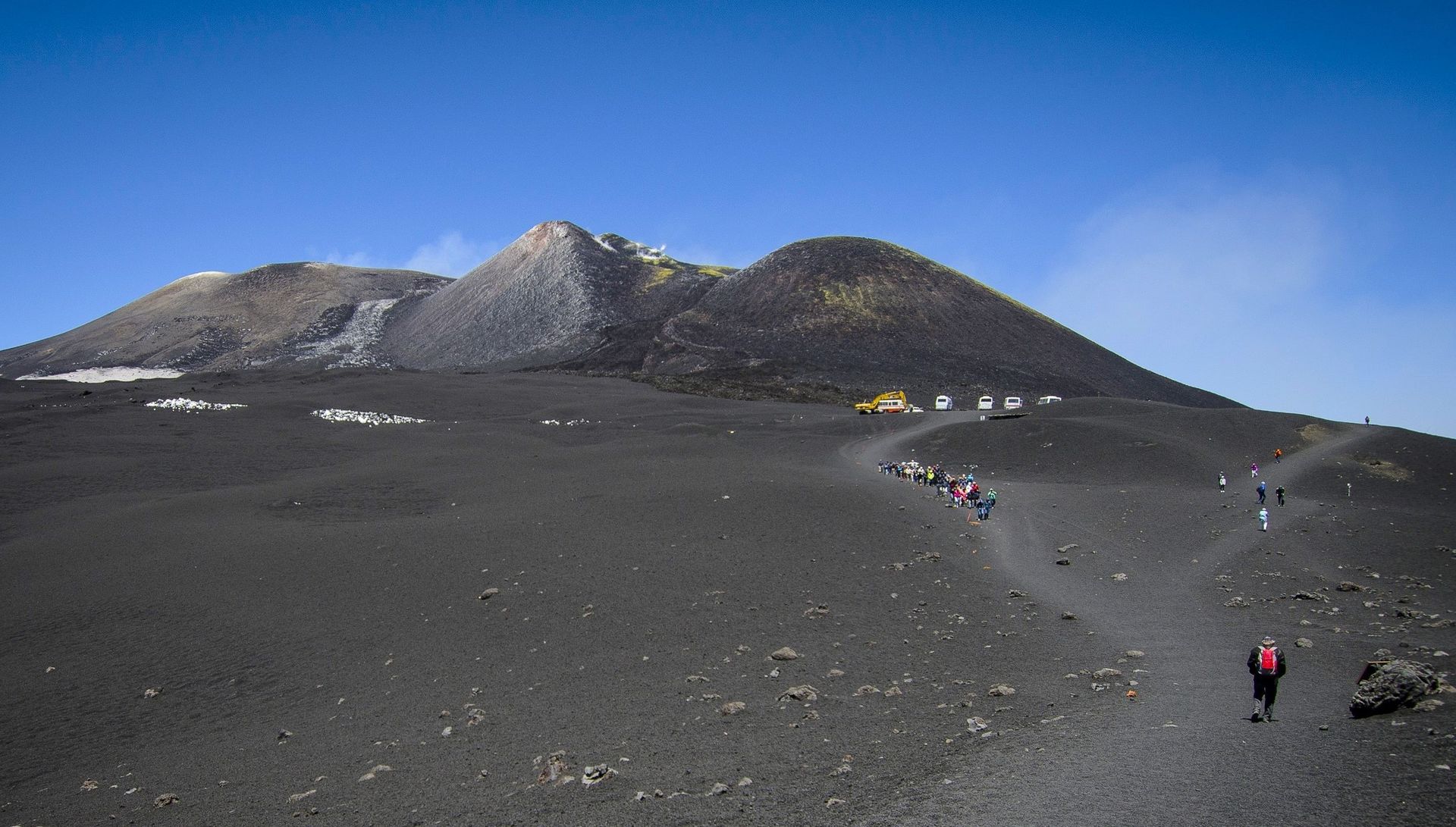 Hikers walking along a rocky volcanic trail toward a dark mountain under a blue sky