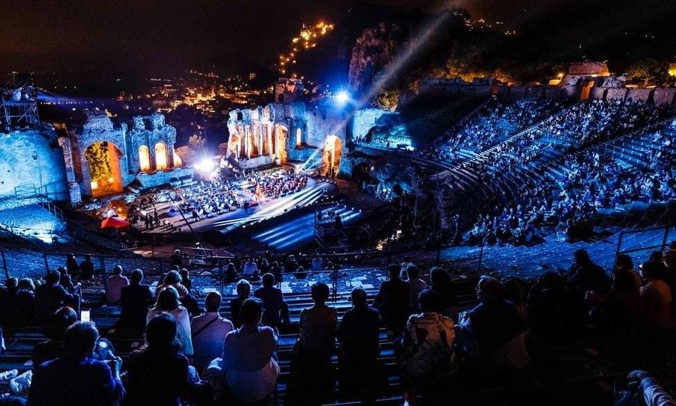 Blue-lit concert arena with a large stage, bright spotlights, and a seated crowd in the foreground