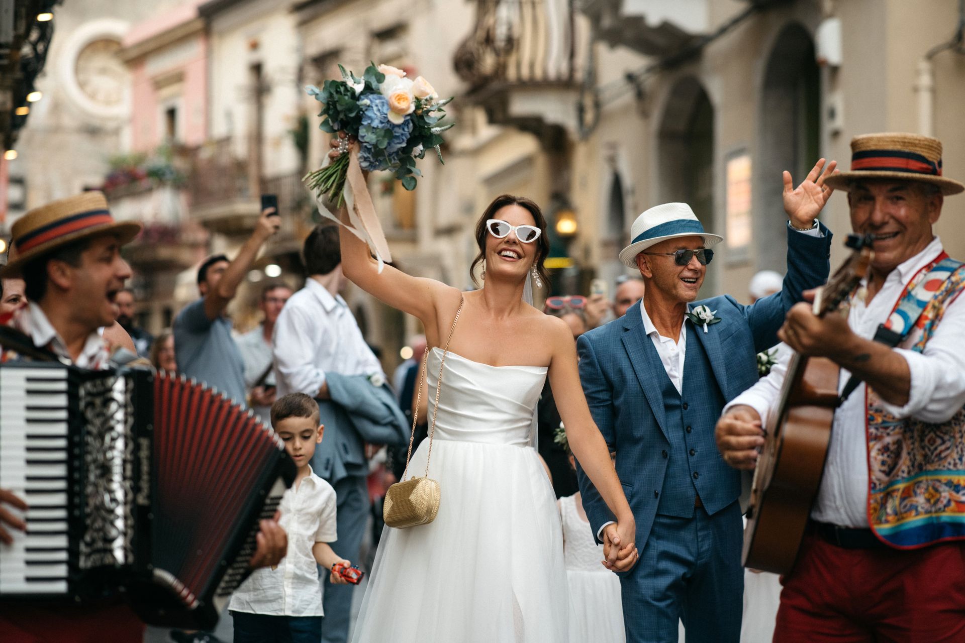 Wedding parade with bride holding bouquet, musicians playing accordion, and guests cheering on a street