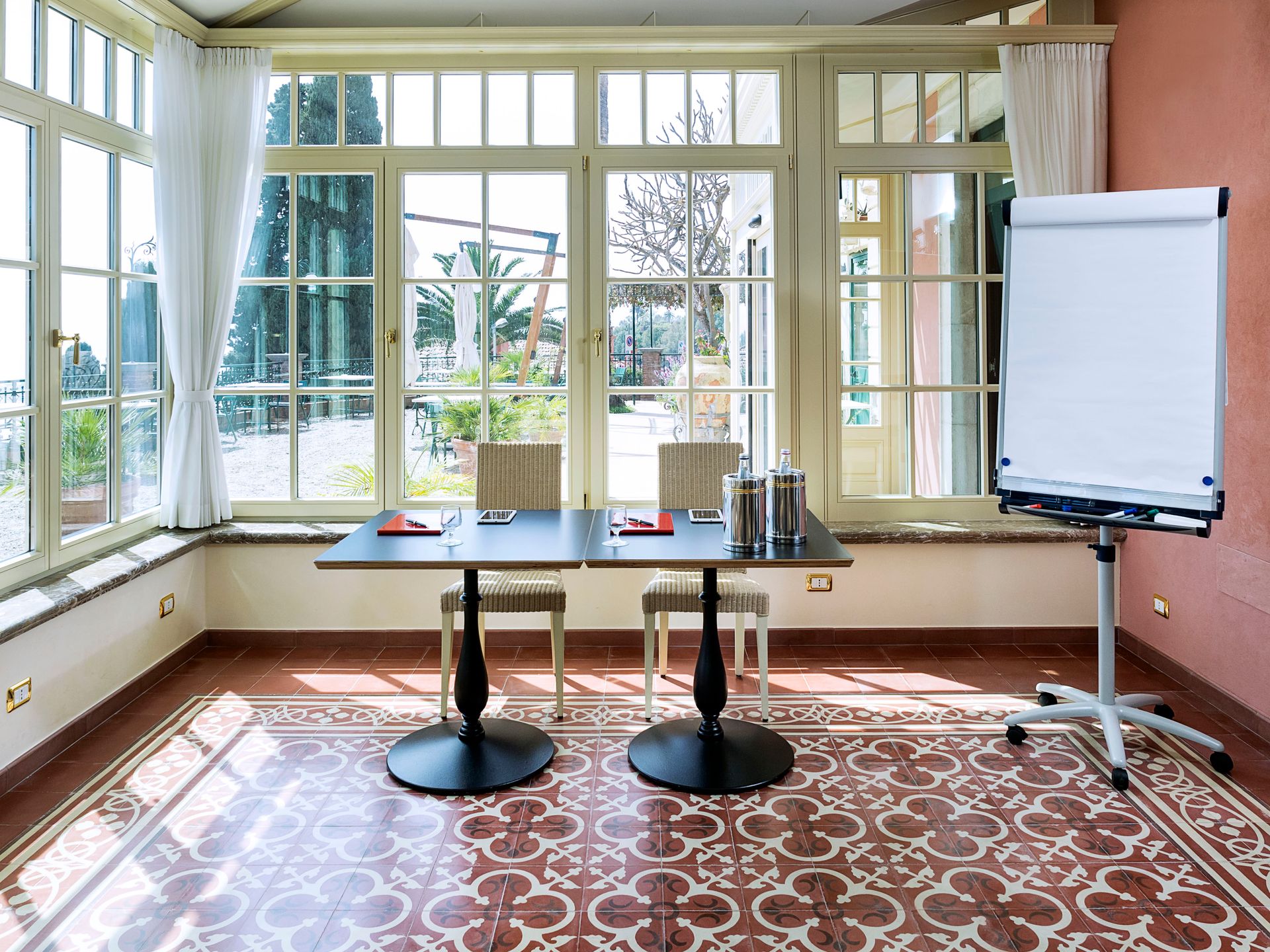 Bright sunlit room with large windows, round tables, and a flip chart easel on a patterned rug.