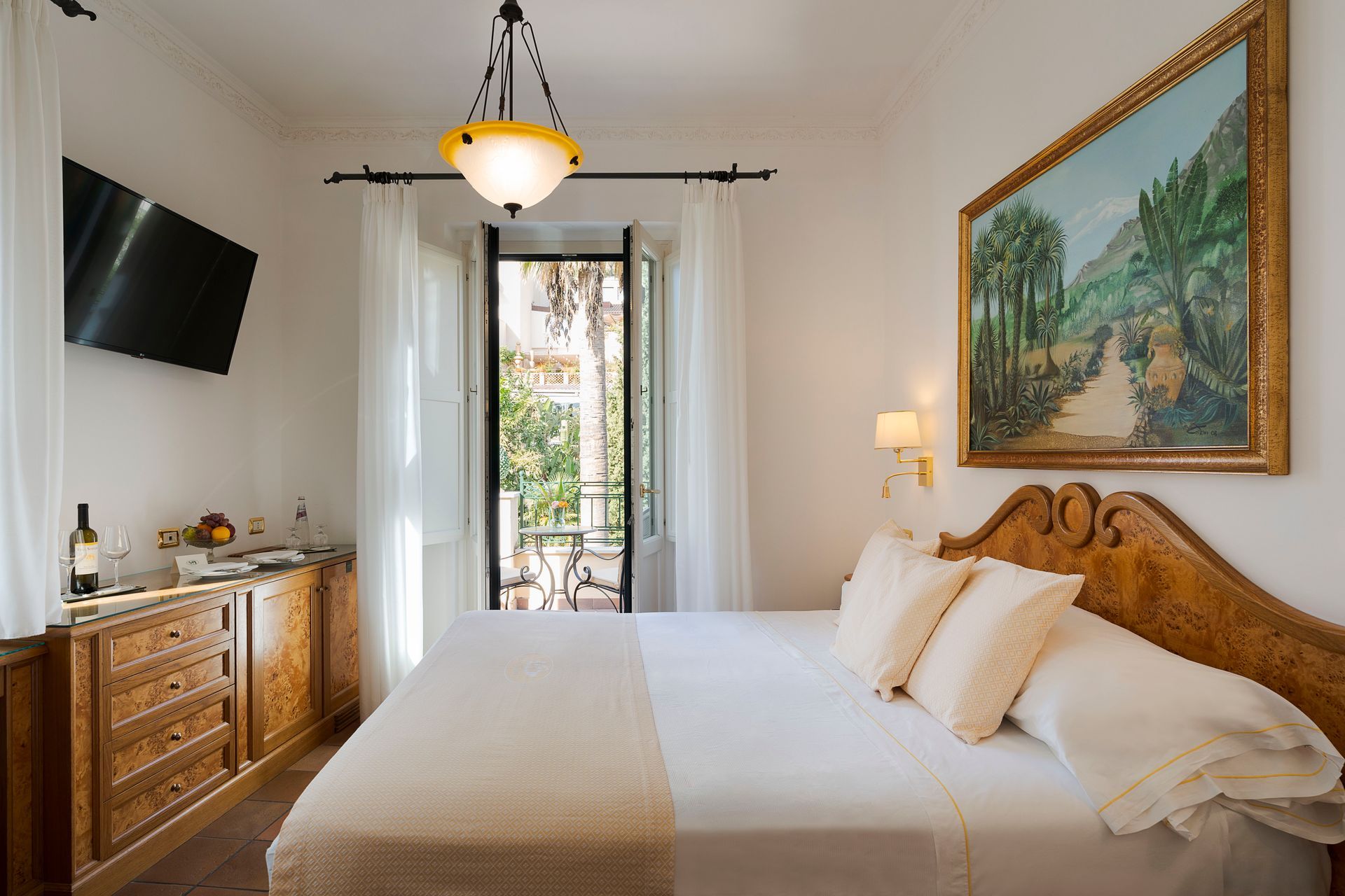 Helles Schlafzimmer mit Bett, Kommode, Wandbildern, Fernseher und Balkontüren, die Tageslicht hereinlassen