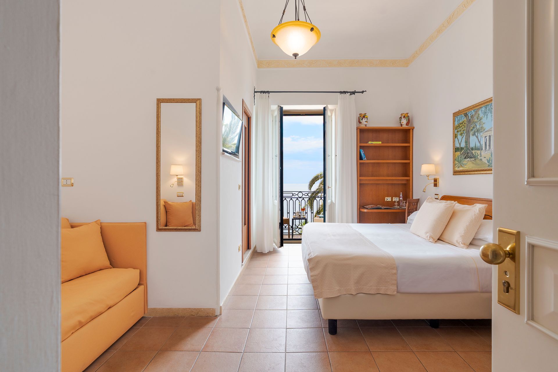 Helles Hotelzimmer mit zwei Einzelbetten, orangefarbenem Sofa, Balkonblick und Holzregalen