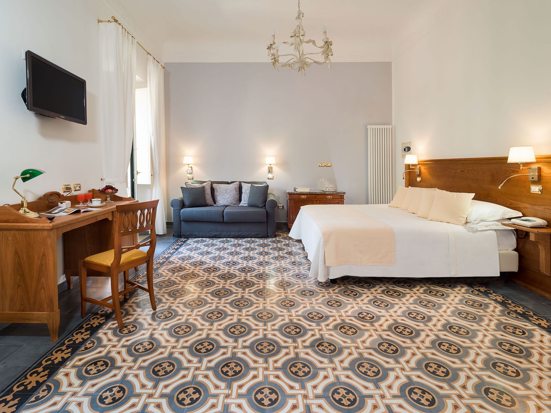 Helles Schlafzimmer mit gemustertem Fliesenboden, Bett, blauem Sofa, Holzschreibtisch und an der Wand montiertem Fernseher