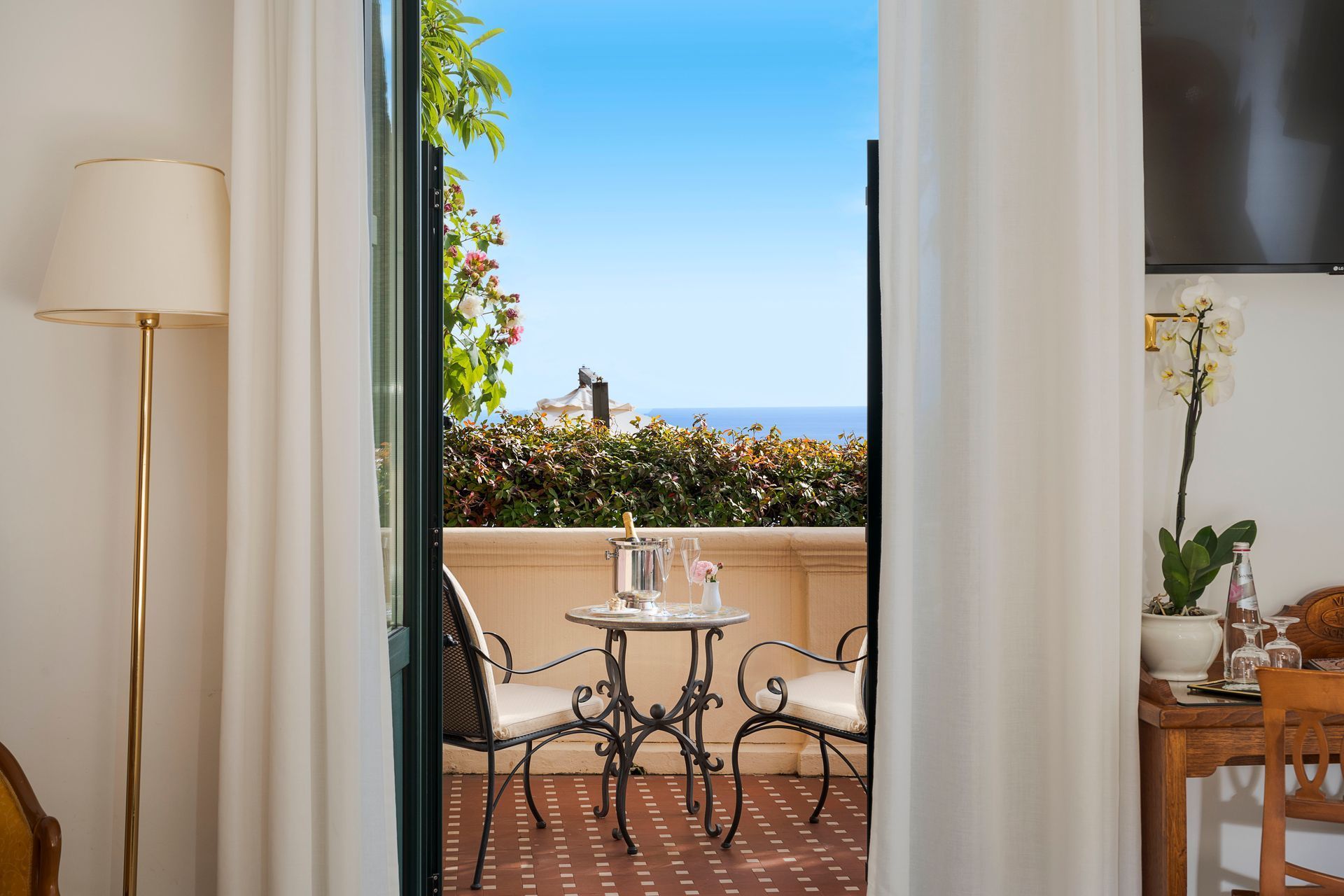 Vue depuis le balcon à travers les rideaux ouverts, avec une petite table, des chaises et la mer bleue