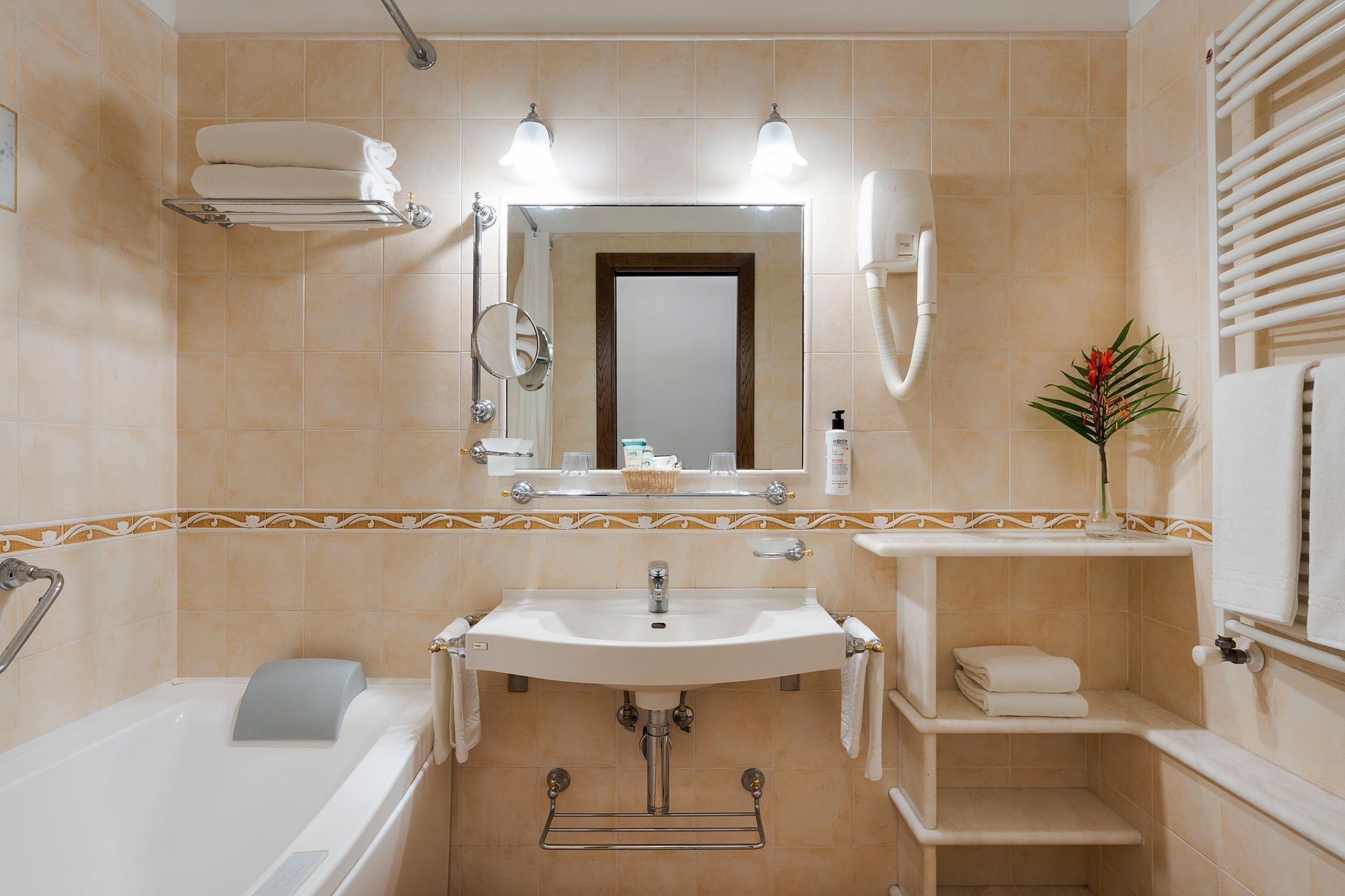 Helles, beigefarbenes Badezimmer mit Badewanne, Waschbecken, großem Spiegel und Wandregalen mit Handtüchern und Dekoration.