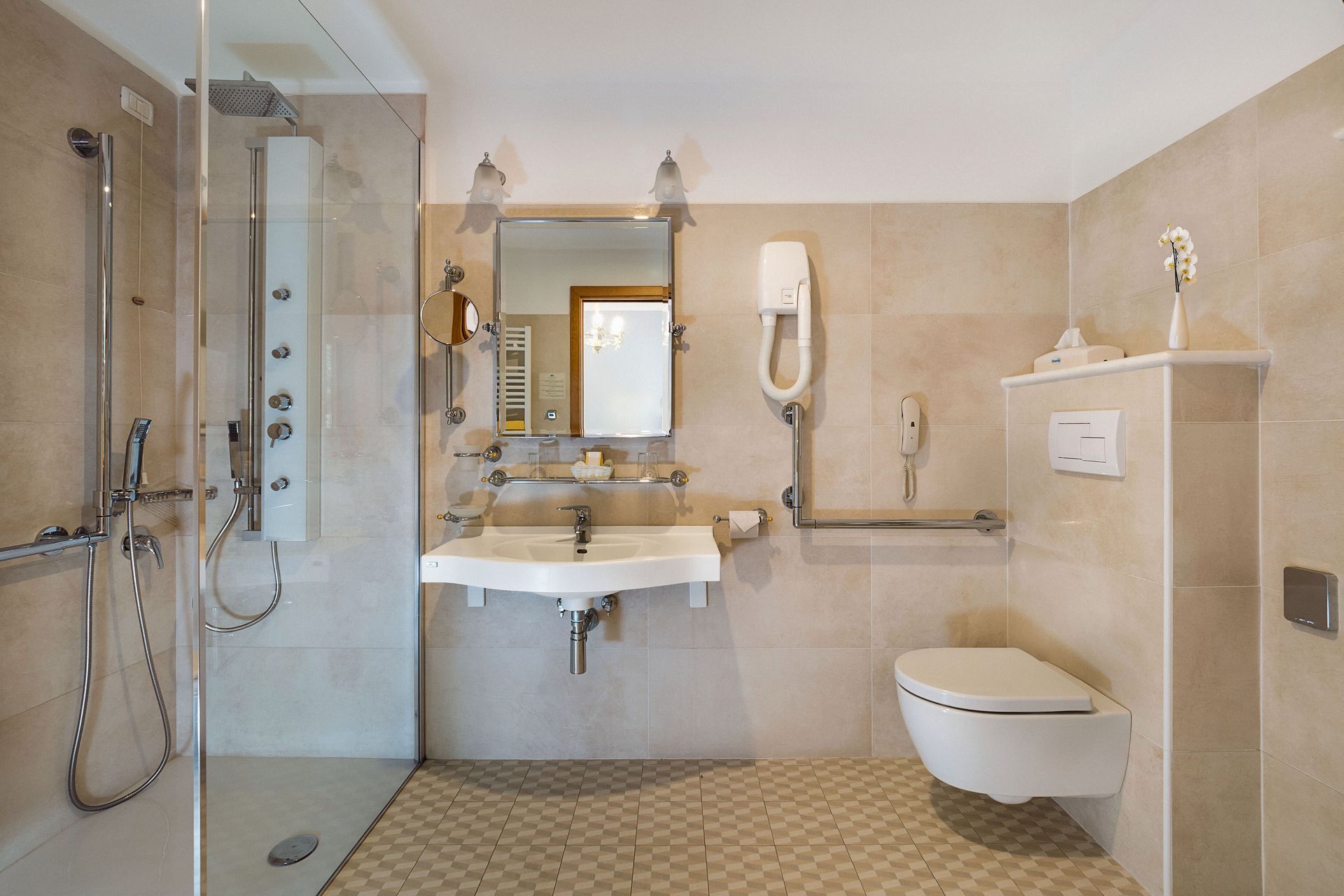 Barrierefreies Badezimmer mit ebenerdiger Dusche, Waschbecken, Haltegriffen und wandhängendem WC in beigefarbenen Fliesen.