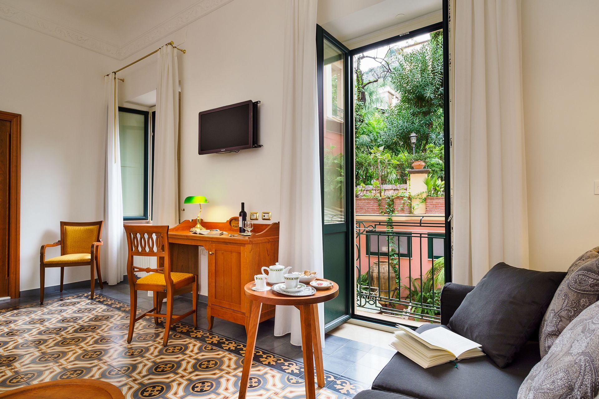 Helles Hotelzimmer mit Schreibtisch, Stühlen, Fernseher, offener Balkontür und einer Couch am Fenster