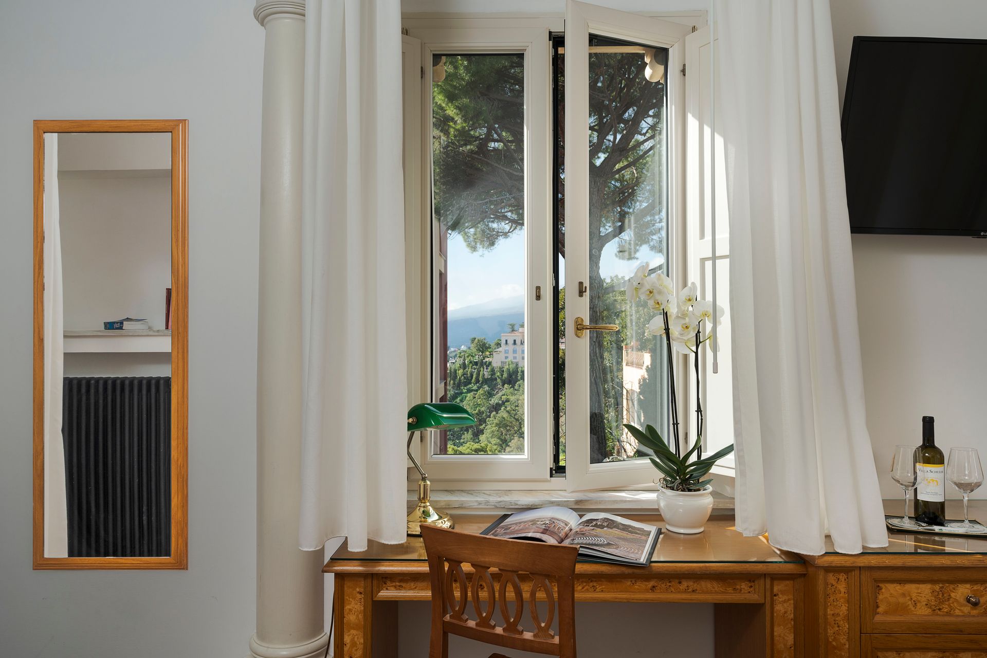 Helles Zimmer mit Holzschreibtisch, Fenster und Vorhängen, Wandspiegel und einem Fernseher auf der rechten Seite.
